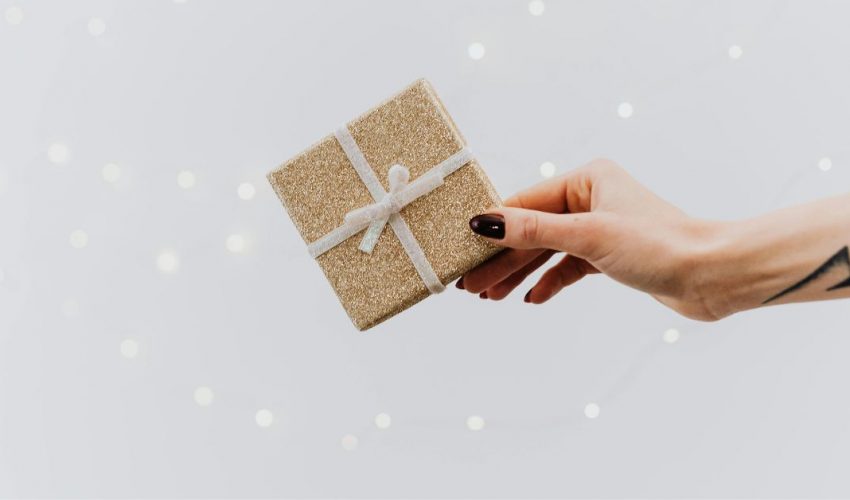 Mano entregando un pequeño regalo de Navidad dorado con lazo blanco. Ideas para regalar experiencias de enoturismo para amantes del vino.